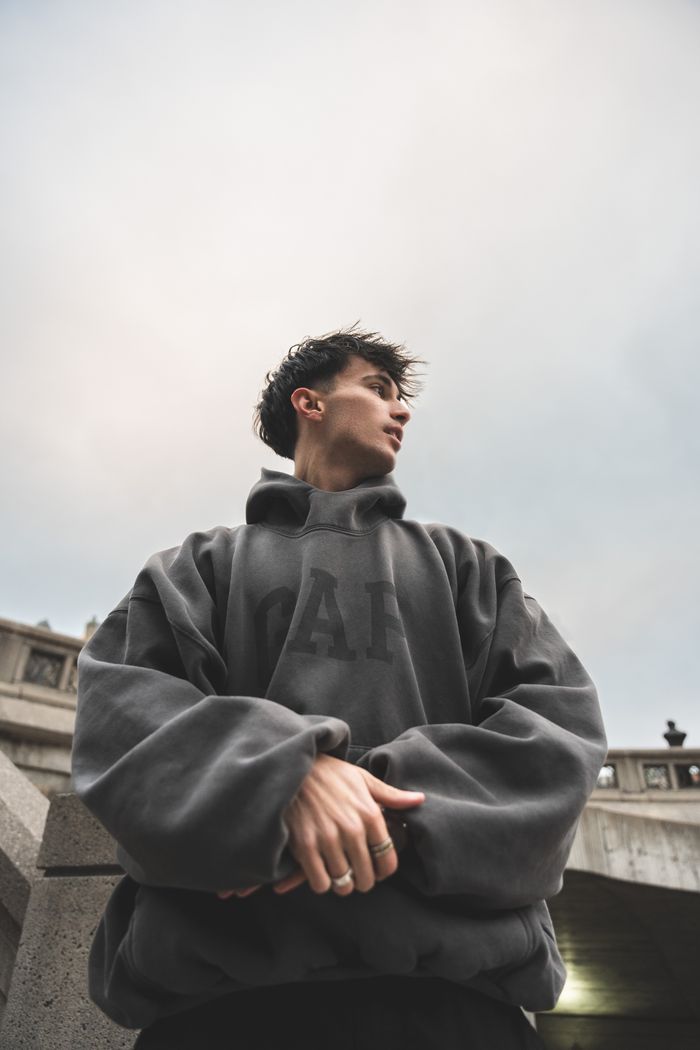 Man in oversized hoodie stands against urban concrete architecture backdrop looking upwards.