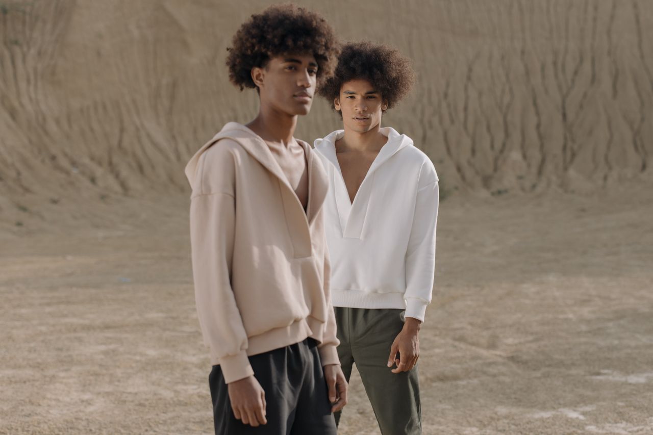 Portrait of two fashionable young men in hoodies posing outdoors with a neutral backdrop.