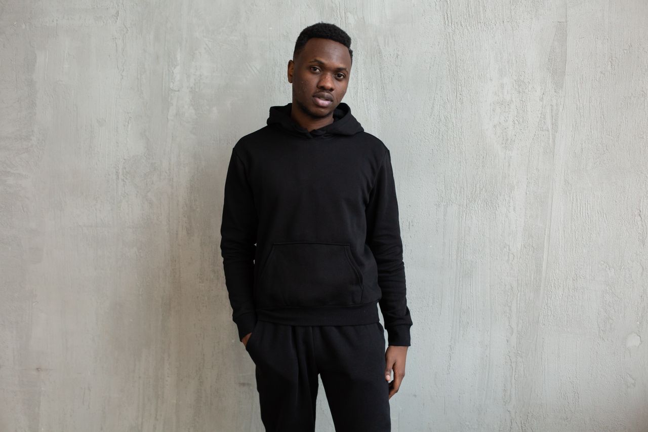 Young unemotional African American male in black comfy hoodie and pants standing with hand in pocket against gray wall and looking at camera calmly