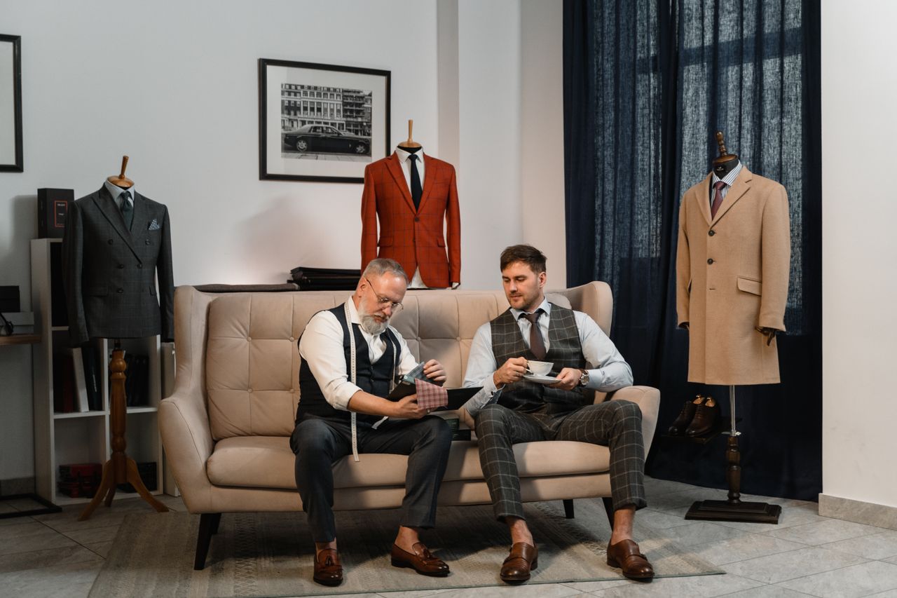 our-mission-01 Two tailors in a stylish workshop discussing custom suit designs surrounded by elegant suits.